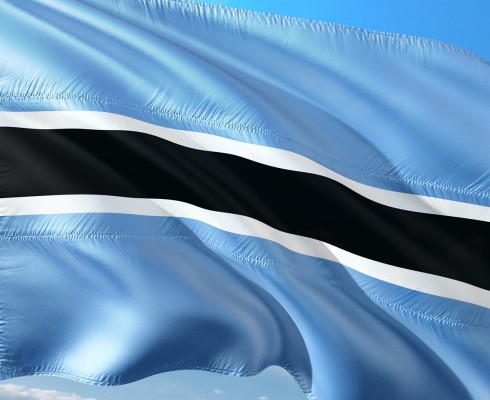 Flagge von Botswana im Wind vor blauem Himmel. Die Flagge ist hellblau mit einem breiten schwarzen Streifen querverlaufend in der Mitte. Der Streifen ist nochmals mit zwei schmalen weißen Streifen abgegrenzt.